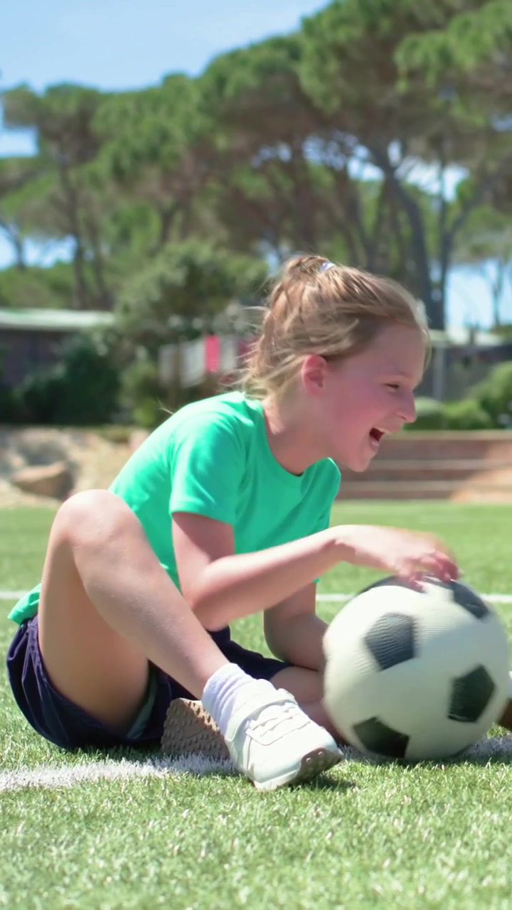 Vídeo vertical: Menina de camisa verde esticando-se para parar bola de futebol rolando na grama sintética, aguardando passe.