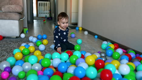 funny adorable toddler comes up to the multiple balls scattered on the floor. kid sits down on his knees and kicking the balls.