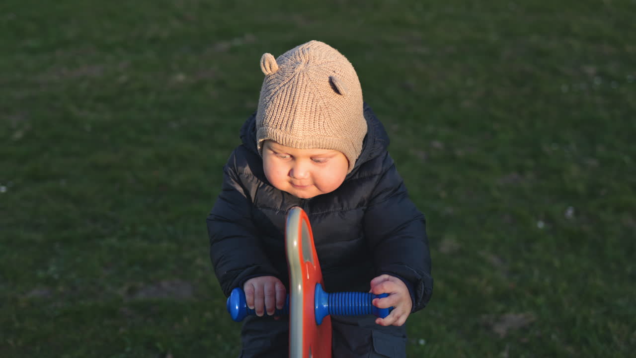 Kleinkind schaukelt auf einem Spielplatz. Ein zweijähriger Junge trägt eine Jacke und Hose und spaziert draußen im Park. Frühling. Kindheit. Glückliches, fröhliches Kind macht seine ersten Schritte.