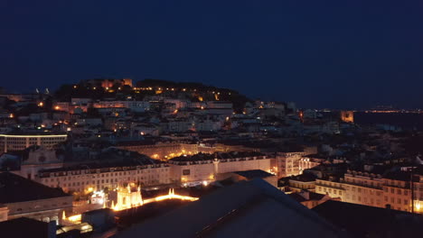 Vista-Aérea-Nocturna-Del-Centro,-Drones-Volando-Sobre-Casas-Y-Plazas.-Castillo-De-San-Jorge-En-La-Colina-De-Fondo.-Lisboa,-Capital-De-Portugal.