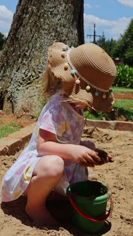 vertical shot of little blonde girl with hat and flower dress playing with mud in sandbox at playground. outdoor playing in sandbox with bucket and mud.