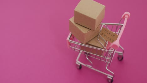 video of black friday text on cardboard and boxes in shopping trolley on pink background