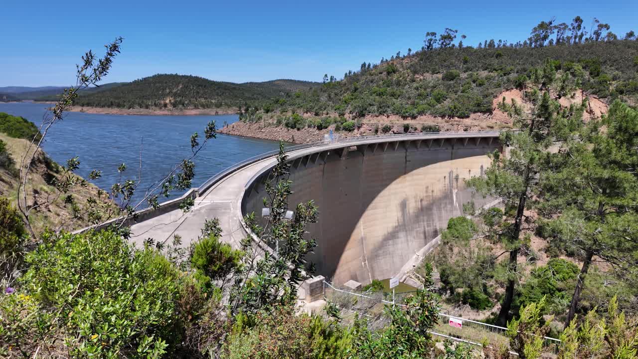 Bravura Dam curving along Odeleite River with surrounding hills and vegetation under a clear blue sky in Portugal