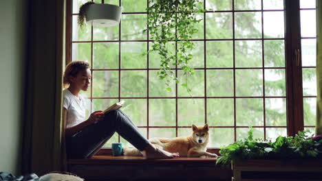 woman reading a book with dog by window
