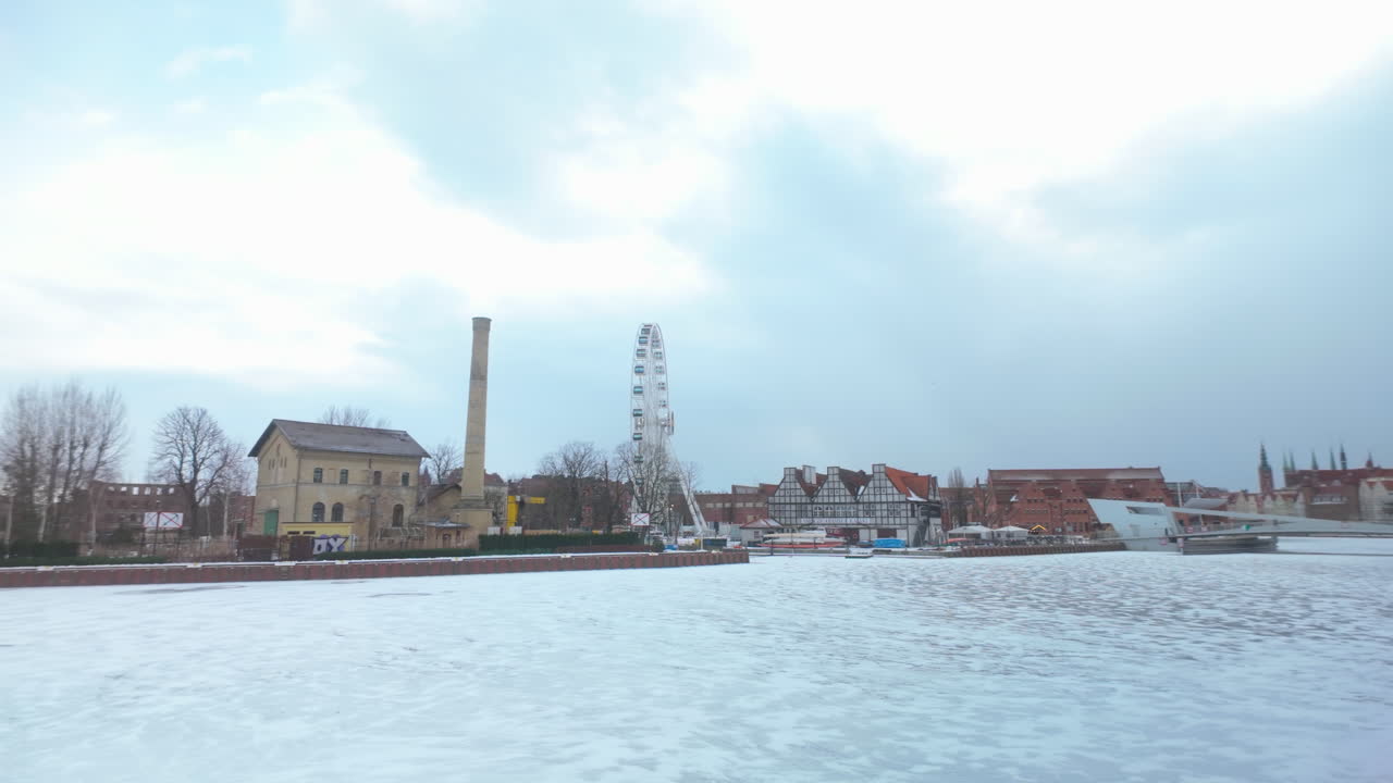 En vidvinkelvy över den historiska strandpromenaden i Gdansks gamla stad på vintern, med den frusna Motlawafloden, pariserhjulet AmberSky och traditionell arkitektur.