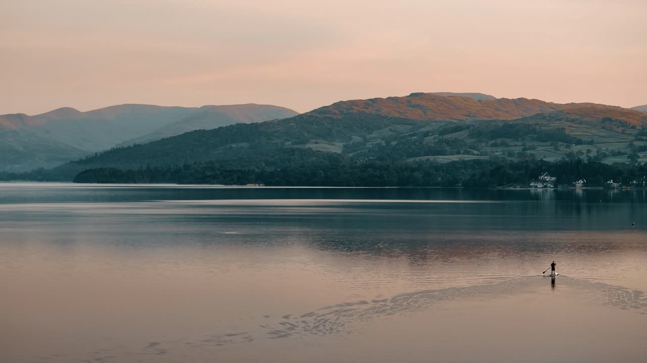 영국의 레이크 디스트릭트 (lake district) 에 있는 윈더미어 호수 (windermere lake) 에 있는 솔로 패들 보더 (solo paddle boarder) 의 공중 도, 산기?? 에 있는 부드러운 오렌지색 알펜글로우 (alpenglow) 와 함께 조용한 물 위에서 멀리 떨어진 곳에 있다.
