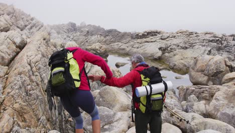 Pareja-De-Excursionistas-Mayores-Con-Mochilas-Y-Bastones-De-Trekking-Tomados-De-La-Mano-Mientras-Escalan-Rocas