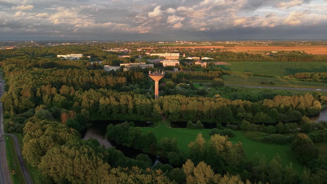 Wideo z drona przedstawia panoramiczne ujęcie z lotu ptaka o zachodzie słońca, ukazujące unikalną wieżę ciśnień wznoszącą się ponad bujnymi zielonymi drzewami, otoczoną spokojnymi stawami i otwartymi polami uprawnymi, z dramatycznymi chmurami i złotym oświetleniem