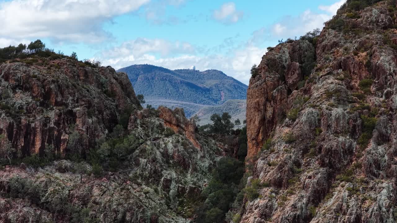 Wide daylight pan reveals rugged Split Rock cliffs, eucalyptus forest, and distant Warrumbungle mountain range under partly cloudy sky in Coonabarabran, Australia