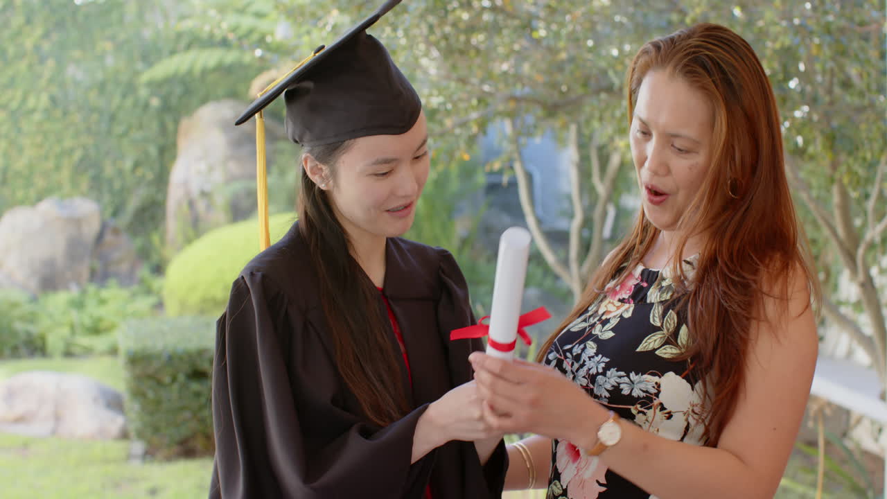 Giovane donna che si laurea, con tocco e toga, che riceve il diploma dalla fiera madre asiatica.