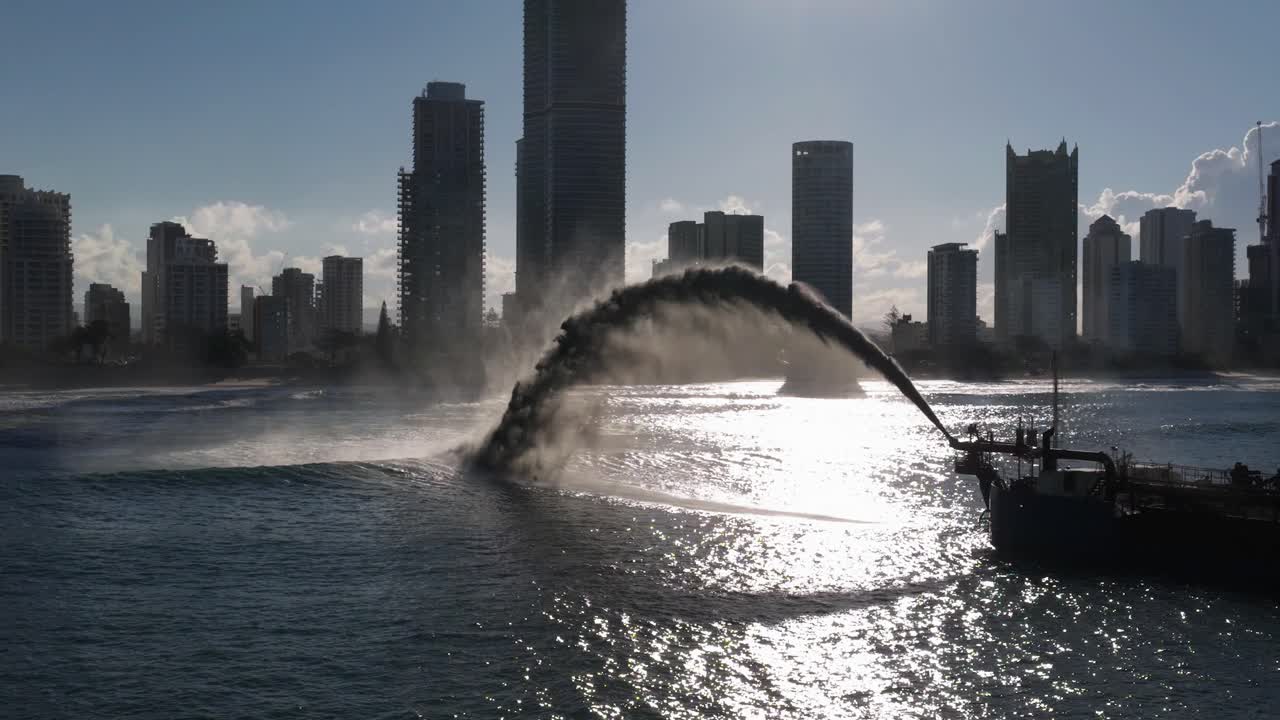 A dredging ship pumps sand to restore a beach, silhouetted against a city skyline under bright sunlight