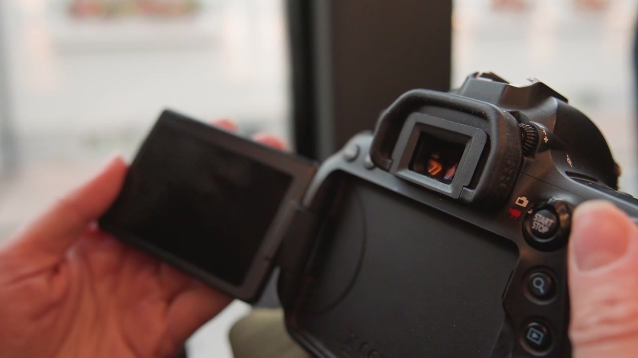 Closeup hands flipping camera screen, content creator testing preview and playback for vlog, checking exposure and focus, stabilizing grip, connecting cable, windowlit indoor setup and steady