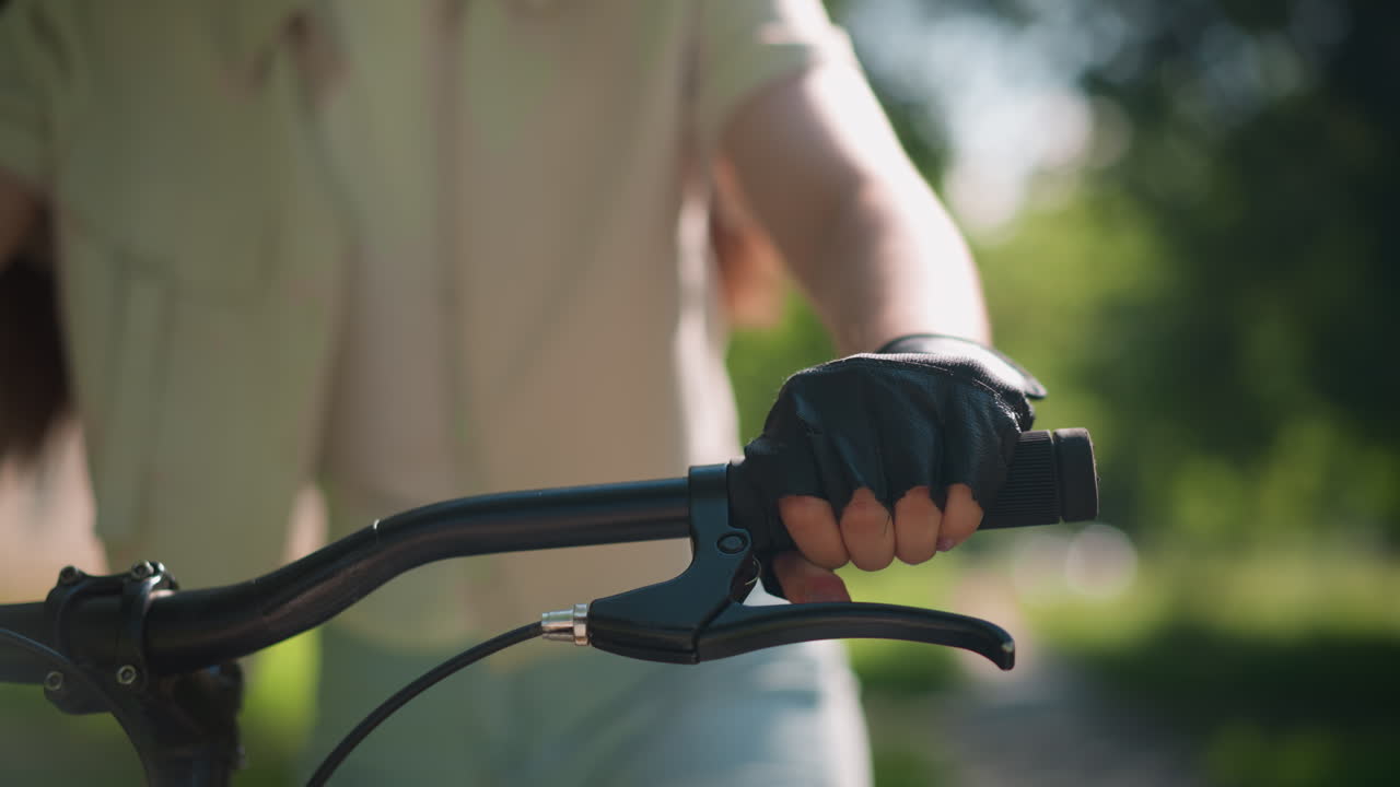 장갑을 입은 손으로 개별적으로 잡는 자전거 손잡이의 클로즈업, 가속화 및 브레이크를 잡고 손에서 반사되는 빛, 배경은 자연 녹색으로 부드럽게 흐릿합니다.