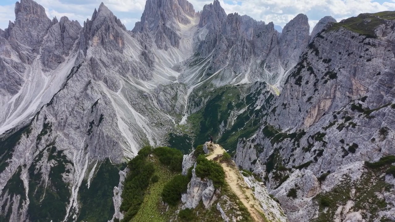 Letecký pohled dronem na ikonickou vyhlídku Cadini di Misurina, s úzkou horskou stezkou obklopenou dramatickými zubatými vápencovými štíty a strmými alpskými útesy v Dolomitech.