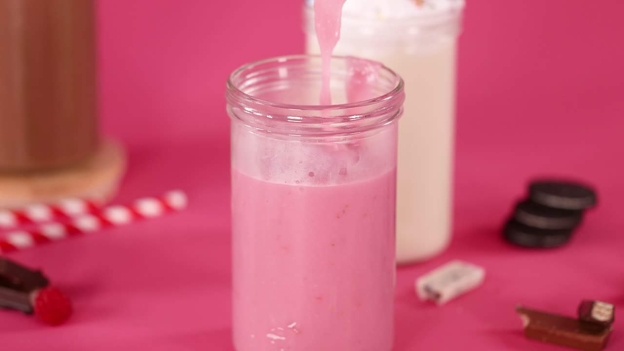 Strawberry milkshake pours into glass, surrounded by cookies, candy, and bright pink background