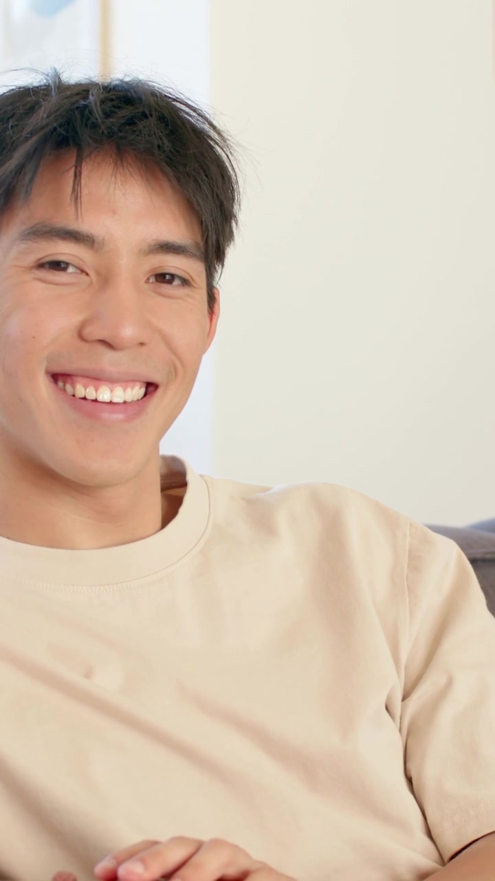 วิดีโอแนวตั้ง: ผู้ชายยิ้มบนโซฟา กำลังตอบสนองต่อสัญญาณจากนอกกล้องที่บ้าน สวมเสื้อสเวตเตอร์สีครีม มีภาพวาดใส่กรอบ
