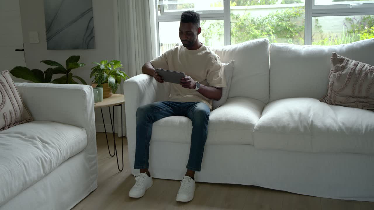 Afroamerikansk mand sidder på en hvid sofa i stuen, holder en tablet og ser et sjovt klip.