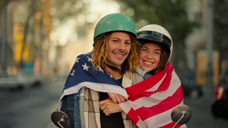 Portrait-of-a-happy-couple-in-helmets-near-a-moped,-a-guy-with-long-curly-hair-hugs-with-his-girlfriend-and-wraps-himself-in-a-US-flag-in-a-summer-city