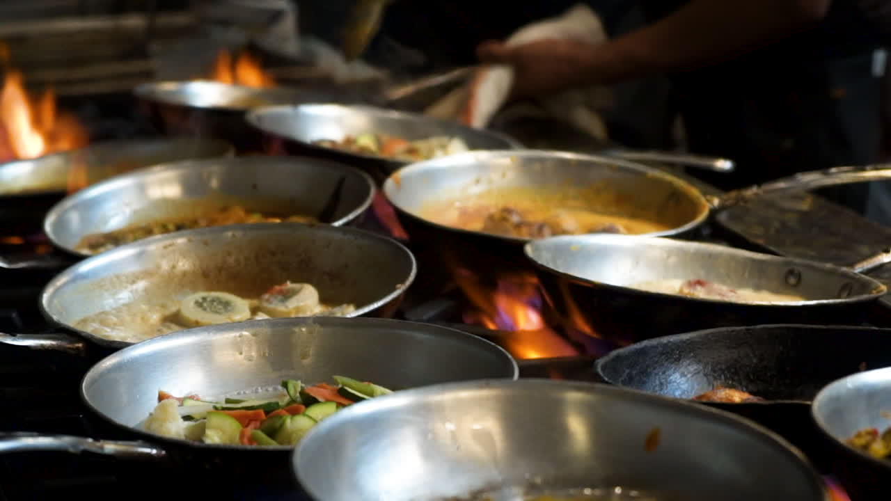 Cinematic view of restaurant stove covered with stainless skillets simmering various menu items, Chef sautes in background, burner flames flare up under pans of bubbling food, slow motion slider HD