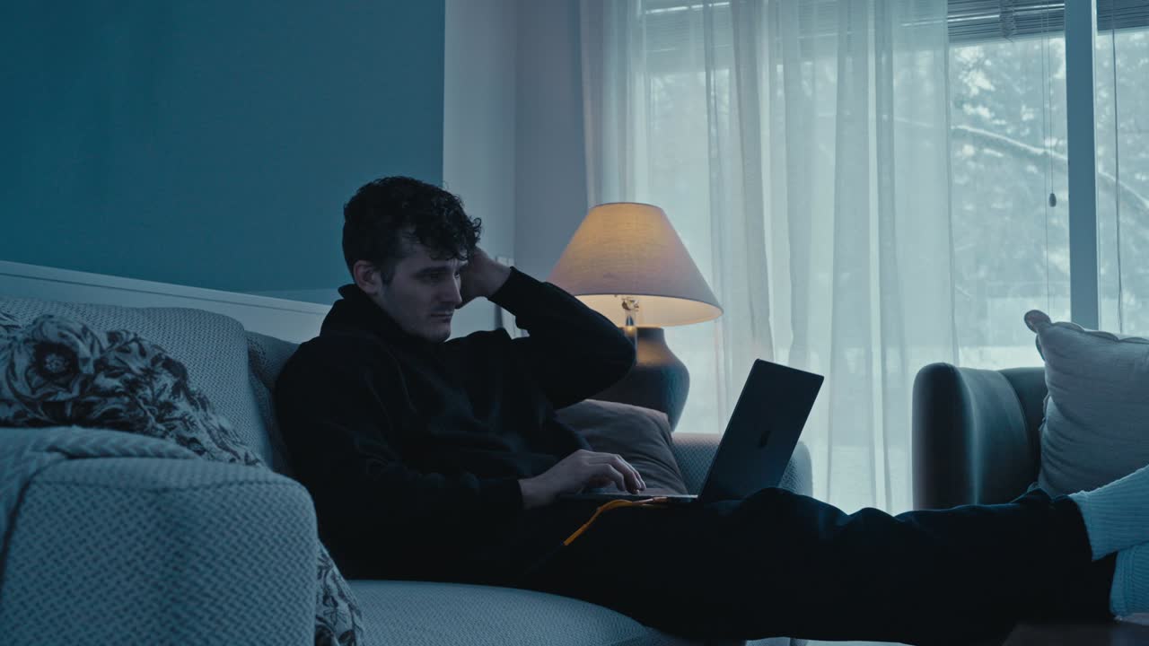 Giovane uomo che usa il laptop sul divano in un salotto in penombra, mentre una bufera di neve e alberi innevati sono visibili fuori dalla finestra.