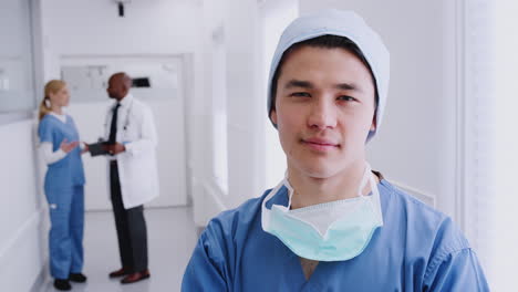 Male-Surgeon-Wearing-Scrubs-And-Mask-Standing-In-Hospital-Corridor-With-Colleagues-In-Background