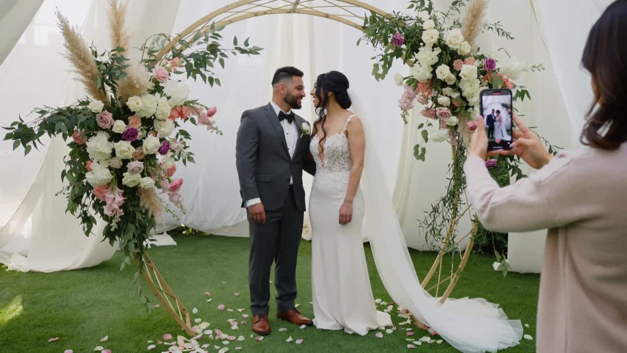 Ospite che alza lo smartphone fotografando una coppia in abiti da sposa sotto un arco dorato, catturando un ritratto