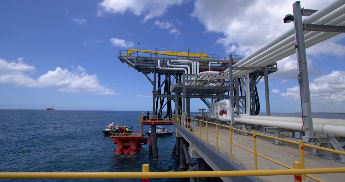 Pull-out tracking shot along pier walkway showing white fuel discharge pipelines and marine unloading platform structure at Coastal Petroleum terminal in San Pedro de Macoris, Dominican Republic