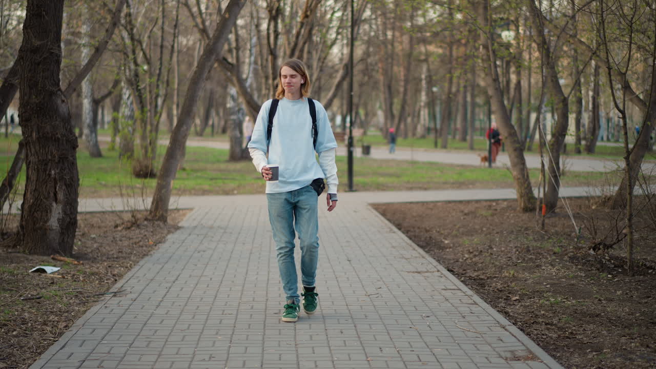 Joven paseando, joven caucásico deambulando solo con una bebida y una mochila, varón caucásico al principio de la edad adulta explorando un parque tranquilo mientras disfruta de un café y lleva una bolsa