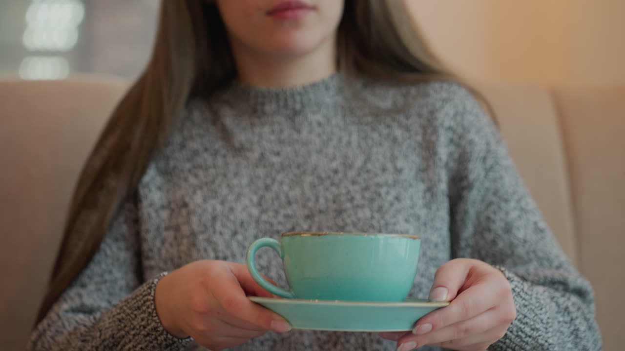 Close-up of a graceful woman placing a hot drink cup on a table after gently sitting down. She appears comfortable in a cozy setting, dressed in a sweater, and taking a moment to relax