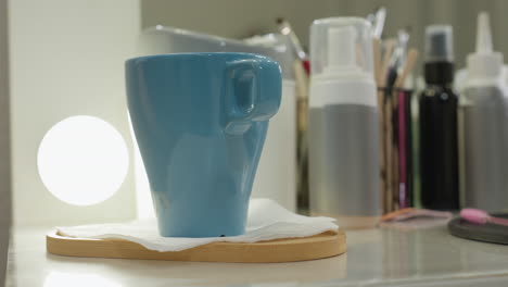 blue ceramic cup placed on wooden tray with tissue on glossy salon table beside glowing light bulb, surrounded by blurred hairdressing tools and products in soft, ambient lighting environment