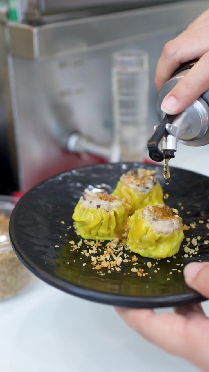 Uma pessoa derrama molho escuro sobre bolinhos shumai amarelos em um prato preto texturizado
