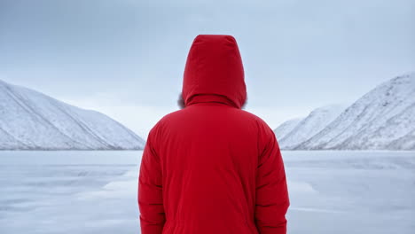 person in red winter coat gazing at a frozen lake and snowy mountains