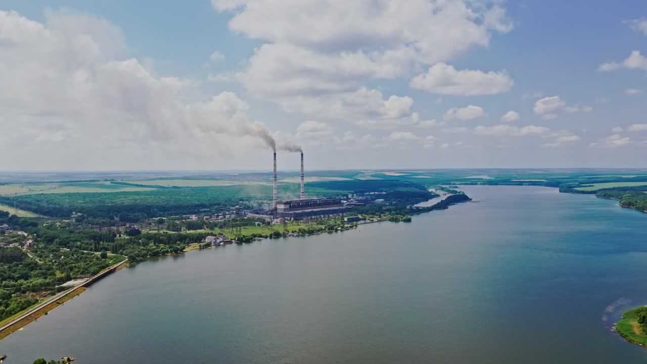 Beautiful scenery of the river background and plant with pipes. Natural landscape of blue water, green fields and factory nearby. Aerial view. Motion up.