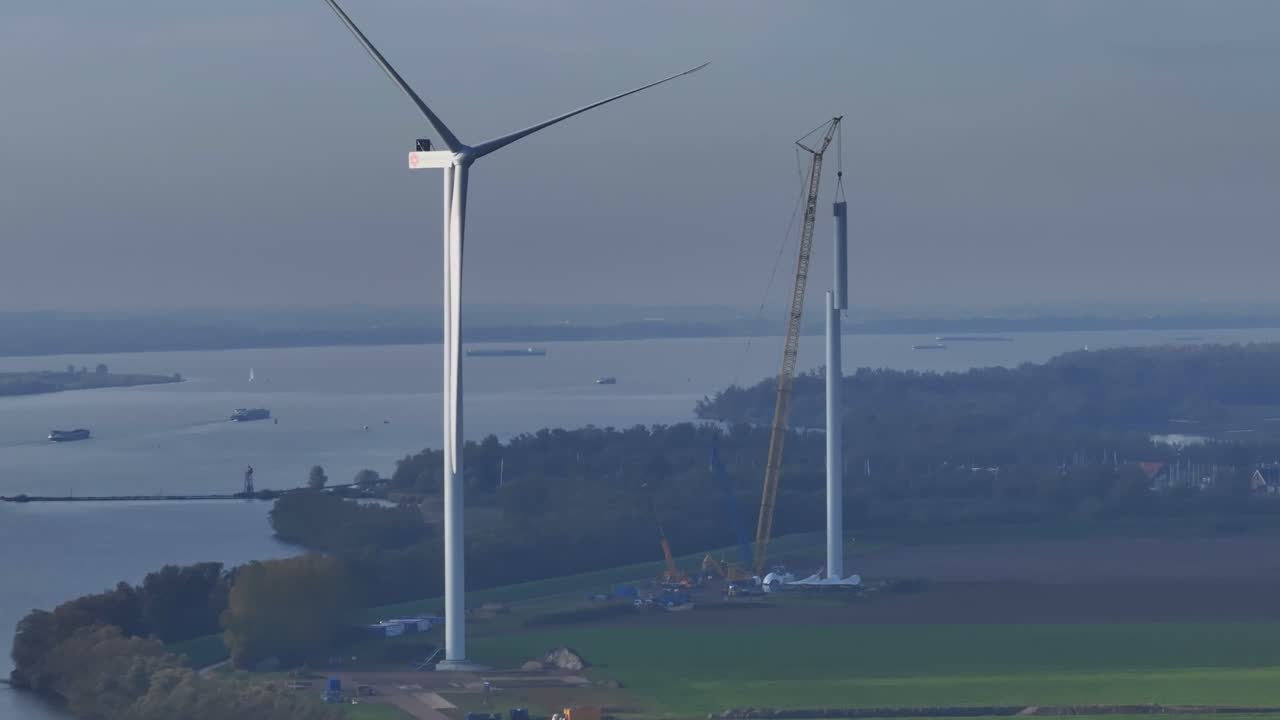 Aerial shot showing two wind turbines, one under construction, with bay and boats in the background.
