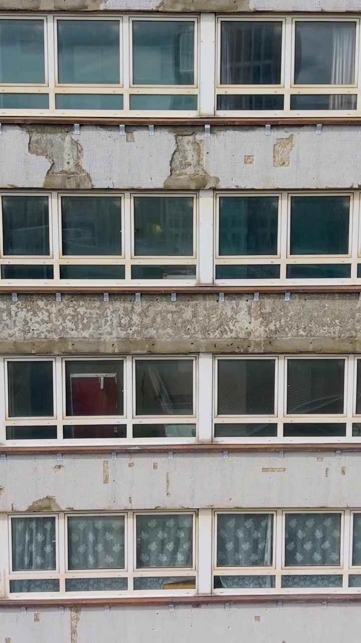 Grattacielo abbandonato a Stratford. Movimento verticale ascendente del drone, rivelando vernice scrostata, intonaco crepato, fasce di cemento macchiate e file di finestre invecchiate su un esterno di grattacielo trascurato