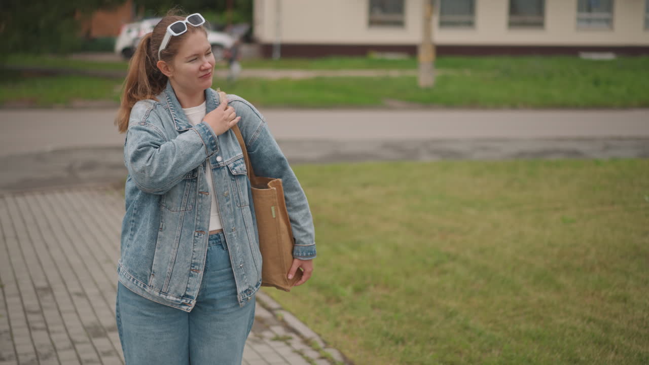 Giovane donna in giacca di jeans che sogna ad occhi aperti su un marciapiede di periferia, occhiali da sole appoggiati sulla testa, tote bag sulla spalla, luce soffusa e nuvolosa, sorriso rilassato, stile street casual, ritratto lifestyle, atmosfera da studentessa