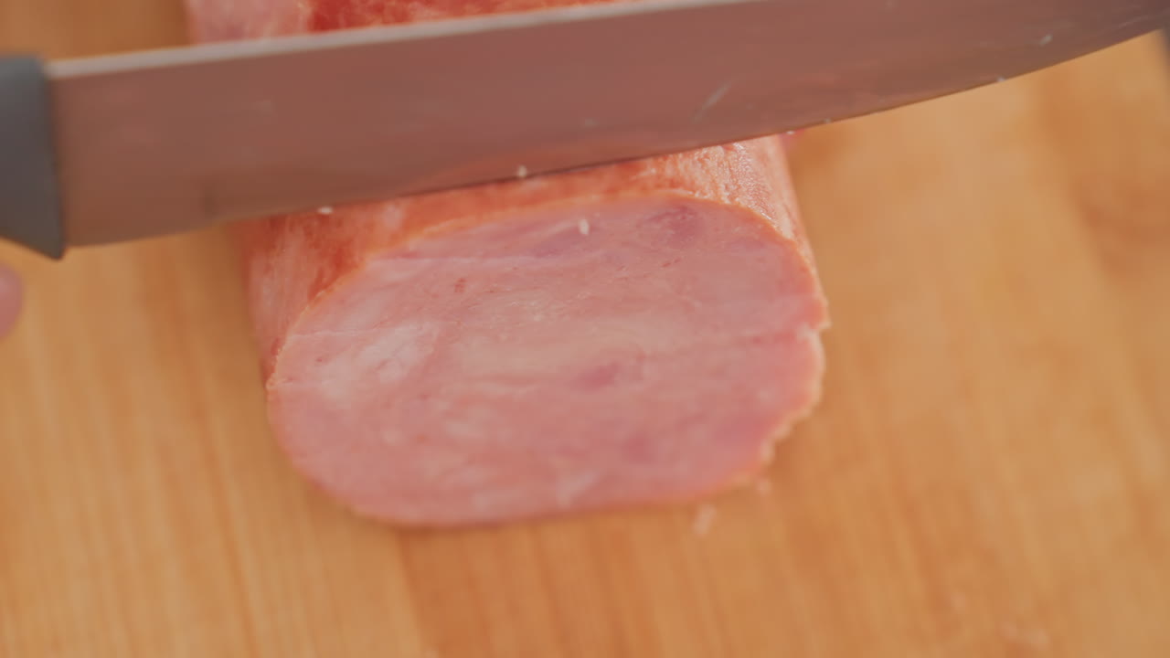 close up top down view person slicing thick round meat on wooden board using sharp kitchen knife in soft blur kitchen setting, showing fine meat texture and precise cutting action in natural light