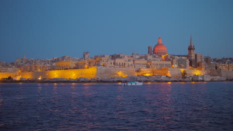 night landscape of valetta skyline in malta with white sailing boat passing. the stunning skyline of old valletta, its churches and towers, its historic remparts from the northern harbour on blue hour
