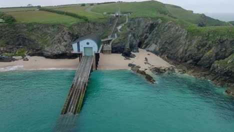 rnli lifeboat station with long slipway descending into clear blue water next to jagged cliffs and sandy beach. camera ascending orbit. mother ivey's bay, cornwall, uk.