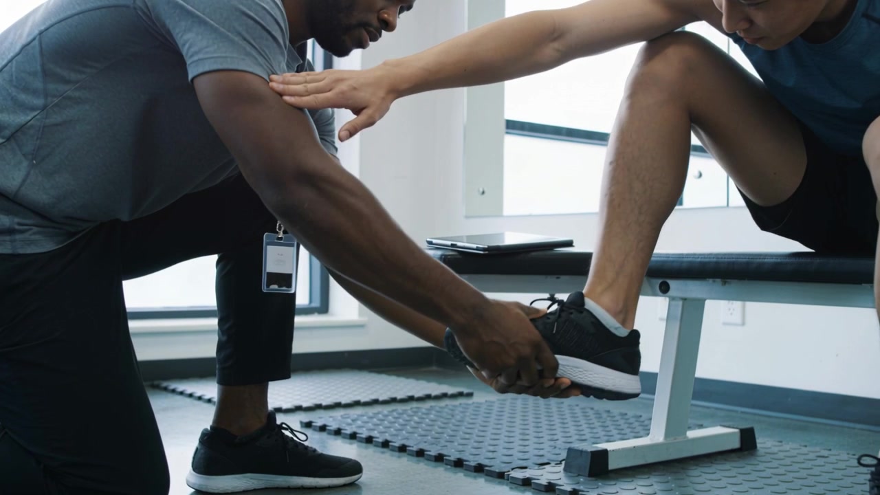 Leaning trainer in grey shirt cupping client's shoe and ankle, guiding dorsiflexion at clinic bench