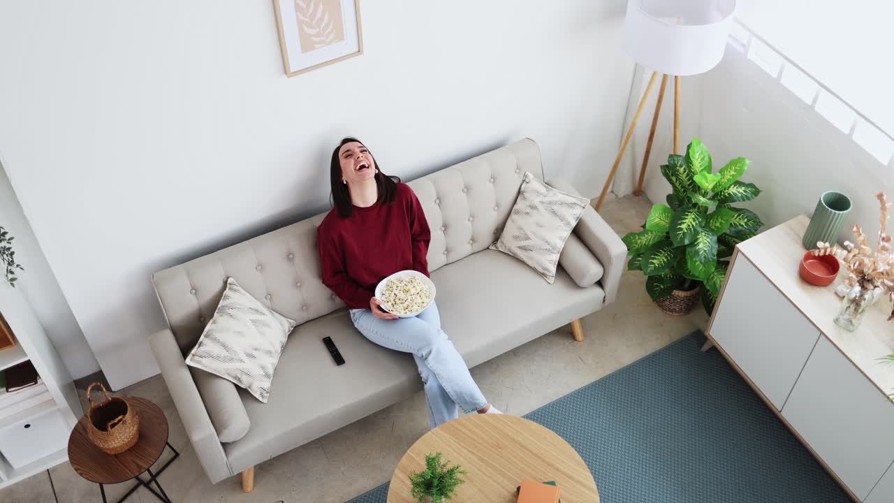 Giovane donna che ride mentre guarda la TV e mangia popcorn sul divano