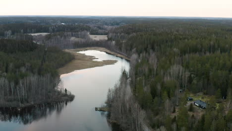 a placid river is seen reflecting the white clouds giving it a bright glow amongst a shoreline covered by a thick forest of ancient green and grey trees.