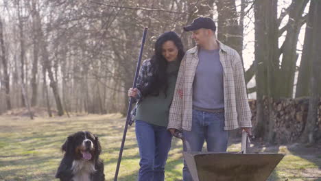 Pareja-Caucásica-Y-Su-Perro-Sosteniendo-Una-Carretilla-Y-Un-Rastrillo-En-El-Campo