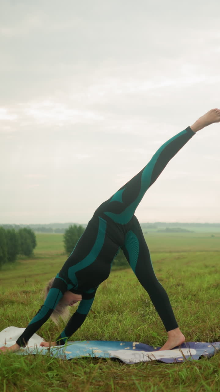 mujer en traje verde y negro practicando yoga dividida de pie al aire libre en una alfombra de yoga en un campo cubierto de hierba bajo un cielo nublado, estirando su cuerpo hacia abajo en un paisaje sereno
