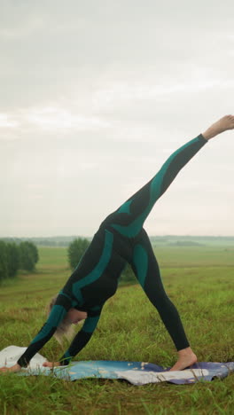 woman in green and black suit practicing standing split yoga pose outdoors on yoga mat in grassy field under cloudy sky, stretching her body downward in a serene landscape