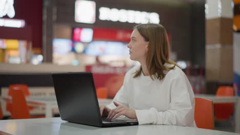 trabajador remoto escribiendo en una computadora portátil en el patio de comidas del centro comercial, sentado en una mesa con un fondo interior moderno con colores vibrantes, señalización de marca y actividad borrosa