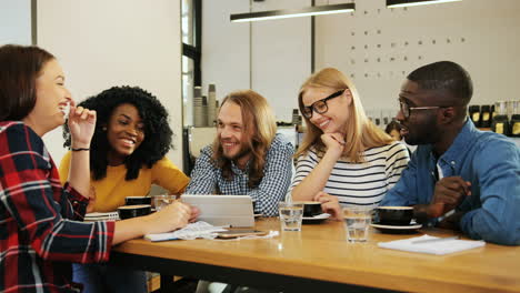 Group-multiethnic-group-of-happy-friends-talking-and-watching-a-video-on-a-tablet-sitting-at-a-table-in-a-cafe