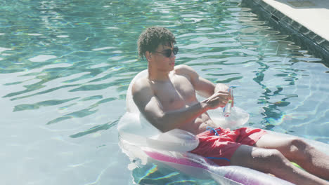 portrait of relaxed biracial man with sunglasses lying on inflatable in swimming pool on sunny day