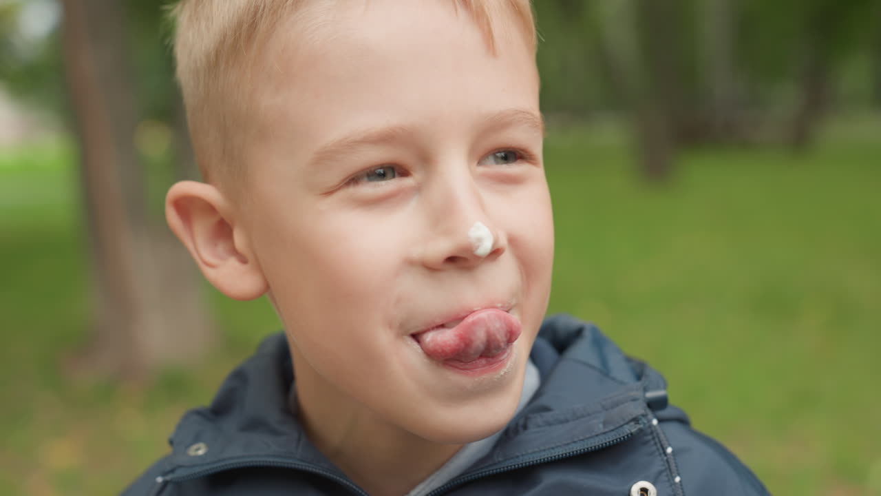 Ragazzo giocoso durante la pausa dopo scuola che lecca un cono con la panna sul naso, sorridente, parco all'aperto, giacca, espressione spensierata, un amico offre un dolcetto, primo piano spontaneo di un momento gioioso dell'infanzia.