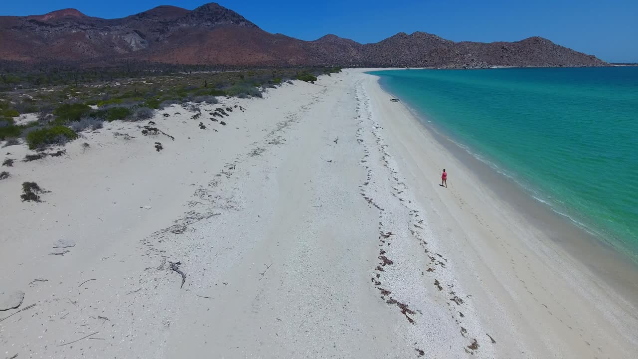 Forward Low Aerial of Girl Walking a Wide White Beach Near Clear Blue Water with Desert Mountain Background